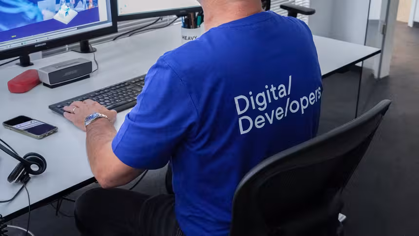 Eine Person mit einem blauen T-Shirt mit der Aufschrift "Digital Developers" sitzt vor einem Monitor 